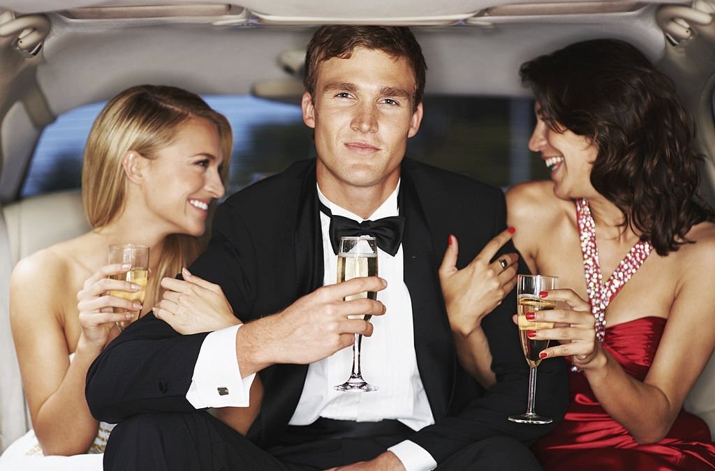 A handsome young celebrity drinking champagne flanked by two gorgeous woman in a limousine